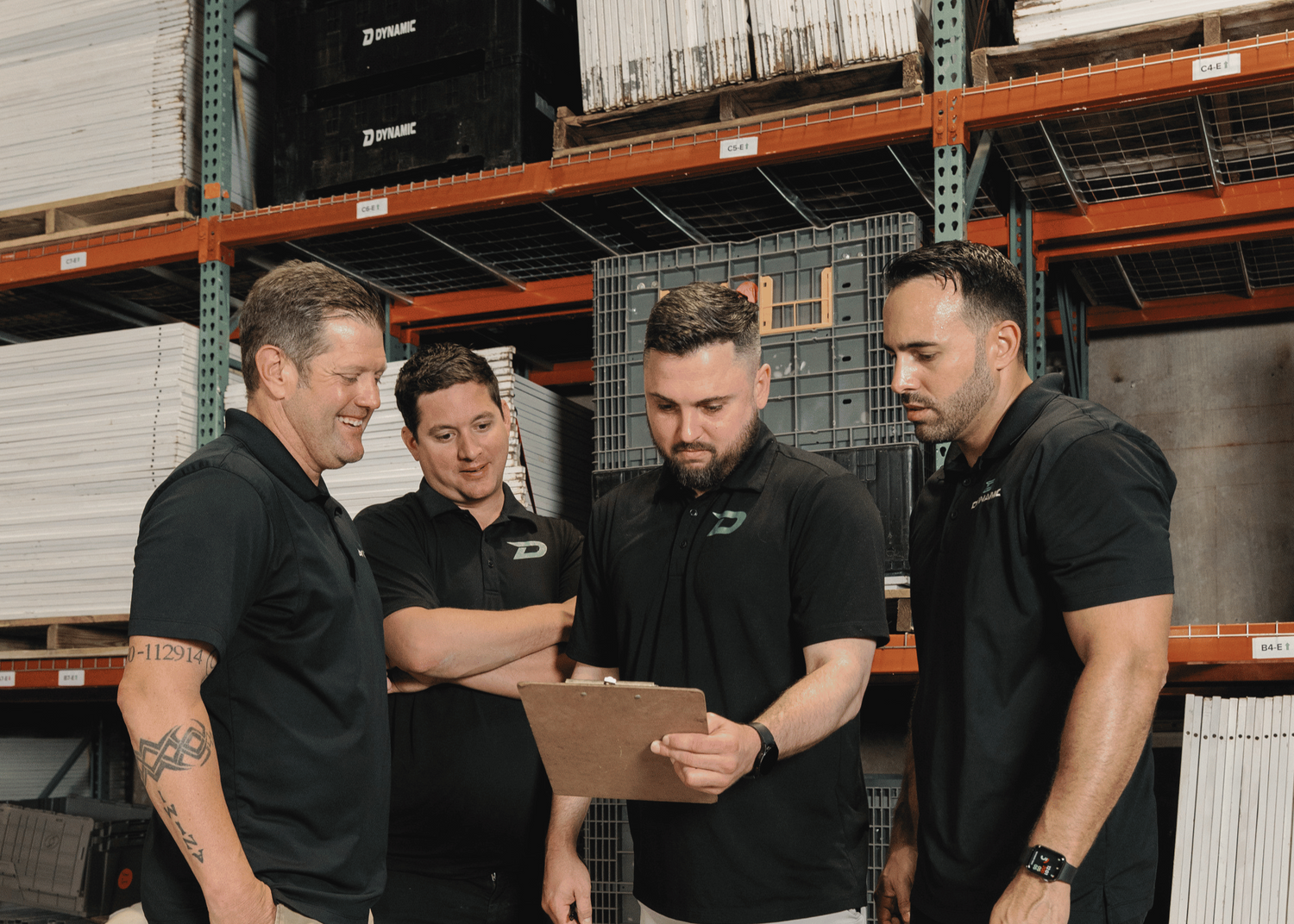 Four men in black polo shirts stand in a warehouse discussing a clipboard document, with shelving units filled with white panels in the background.