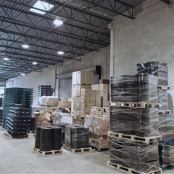 A large warehouse interior with stacks of pallets, some wrapped in clear plastic, and cardboard boxes under a high industrial ceiling with exposed metal trusses and overhead lights.