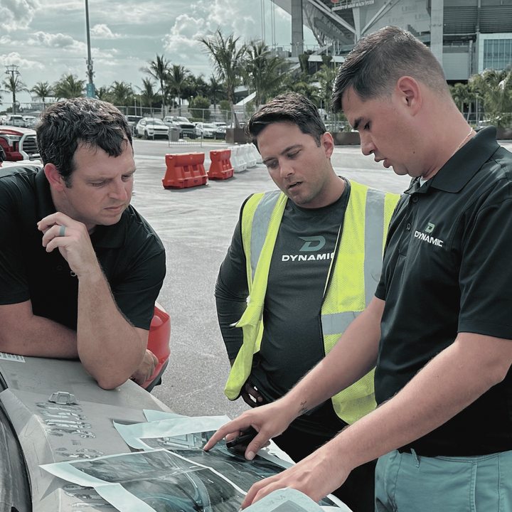 Three men standing outdoors near a car, reviewing a printed map or document. One man wears a bright yellow safety vest over a dark shirt, and the setting includes a parking lot, palm trees, and a large modern building in the background.