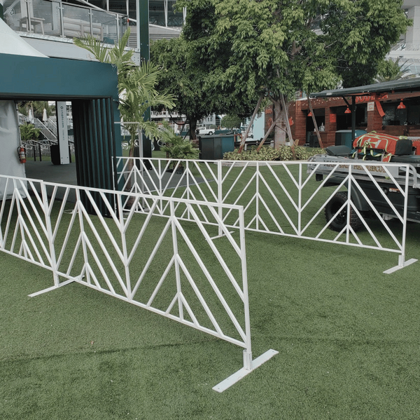 White metal event barricades with a geometric chevron pattern arranged on green artificial turf, with a teal tent structure and outdoor venue background.